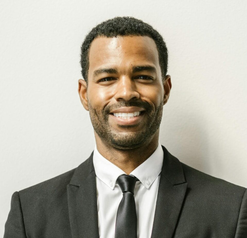 Confident man in formal attire smiling against a plain white wall.